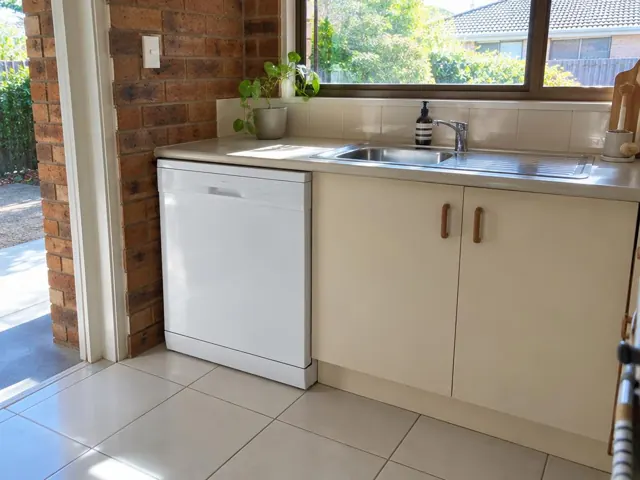 White dishwasher installed under a kitchen bench beside a sink, with brick wall and tiled floor in Edgeworth